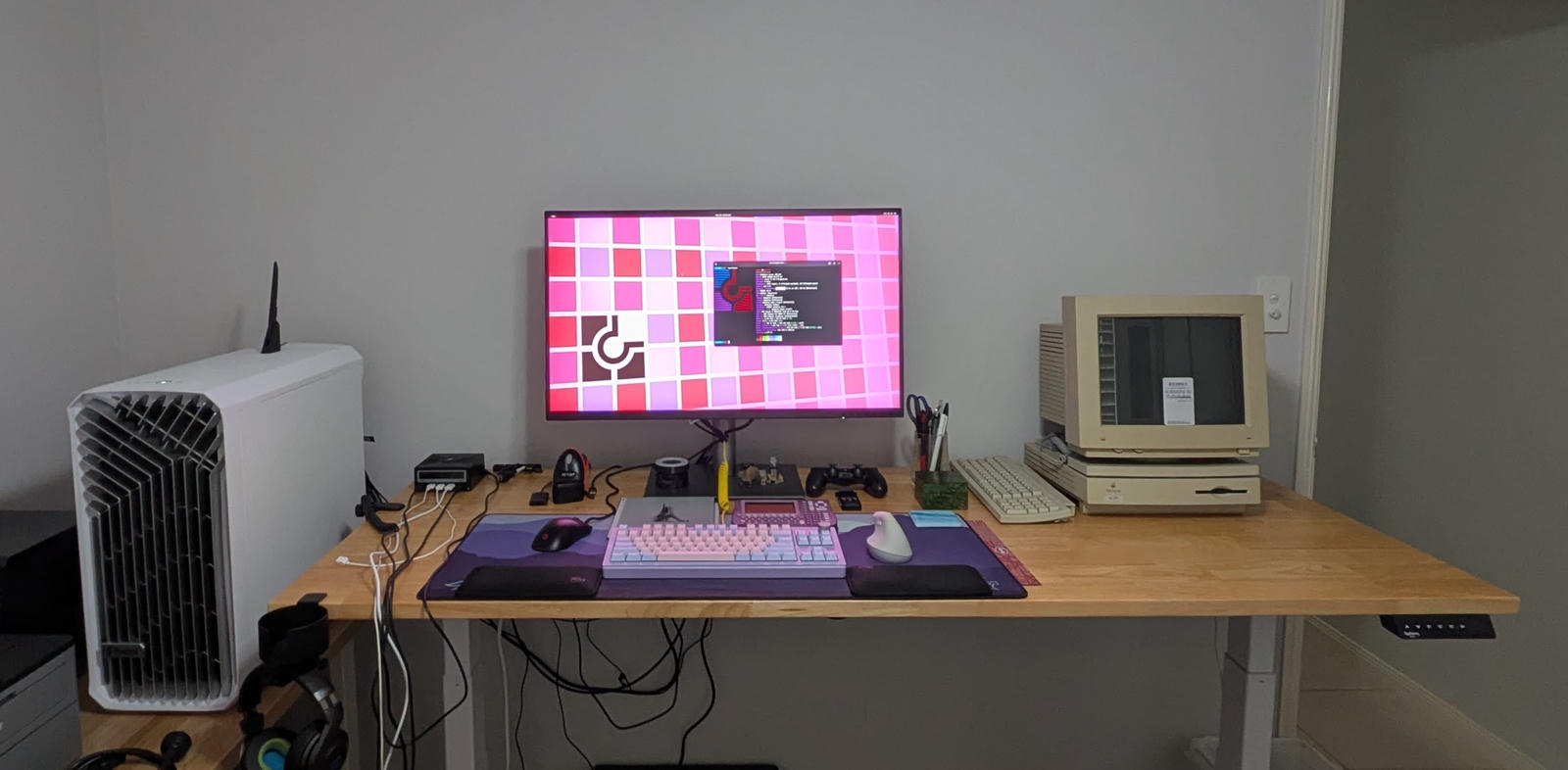 A photo of my standing desk. There's a large display showing a Chimera Linux GNOME desktop, on the right is Macintosh LC 475. In front of the monitor is: a PS4 controller, TI voyage 200 graphing calculator, tenkeyless mechanical keyboard, mouse, and Boox Go 103 e-Note.