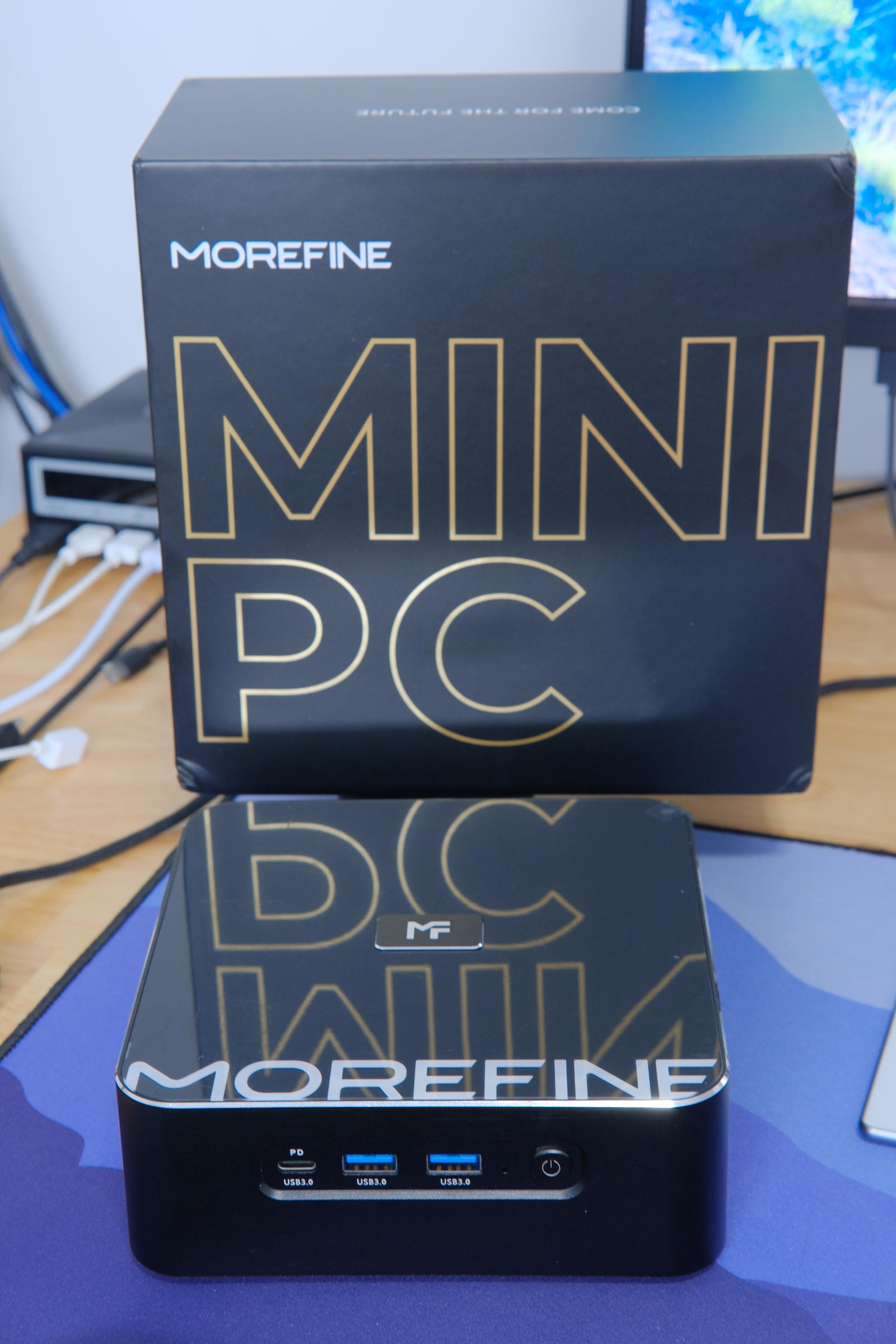 Photo of the MOREFINE mini PC with its box in the background. The box has the text 'MINI PC' in large letters outlined in gold, which is reflecting in the top of the PC.