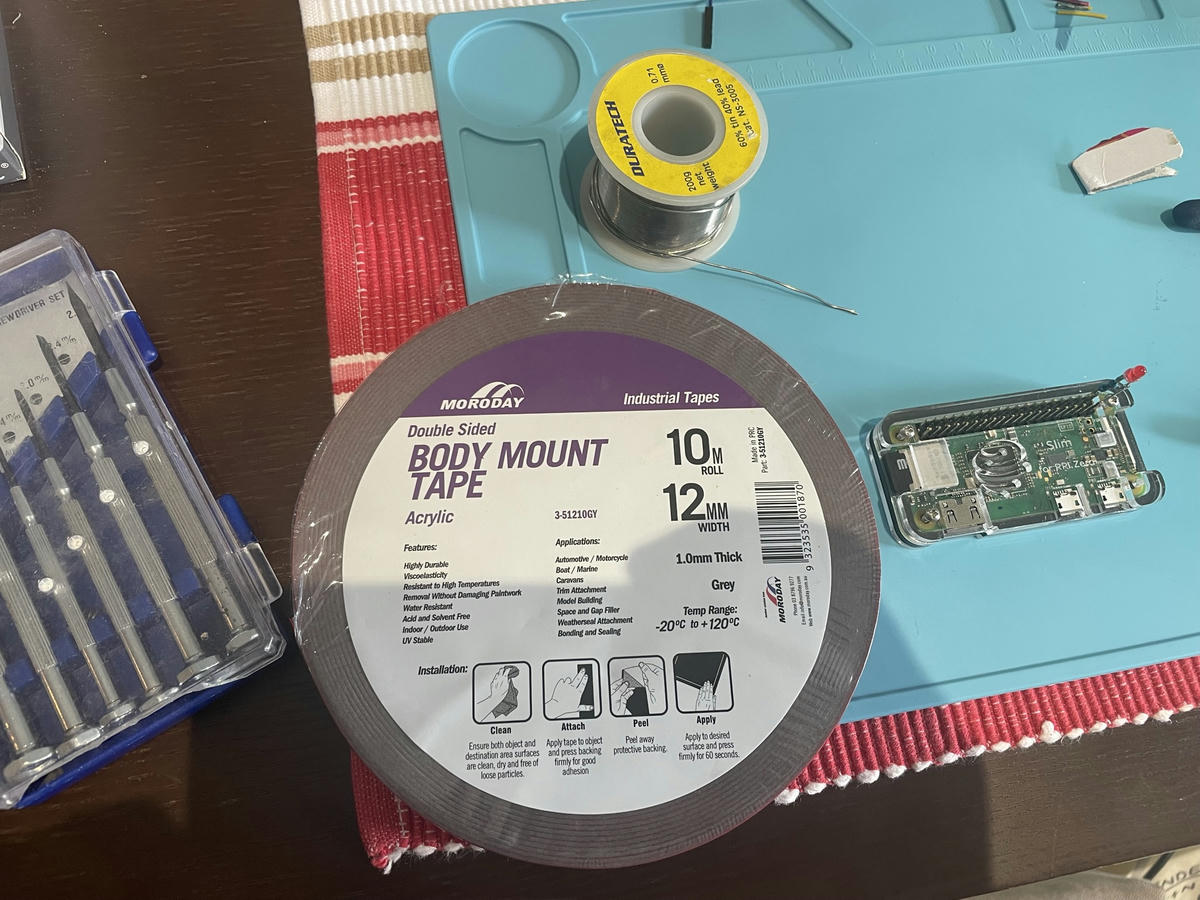 Photo of a roll of double sided body mounting tape on a table with mini screwdrivers to the left, a roll of solder above, and a Raspberry Pi Zero with an LED attached to the right.