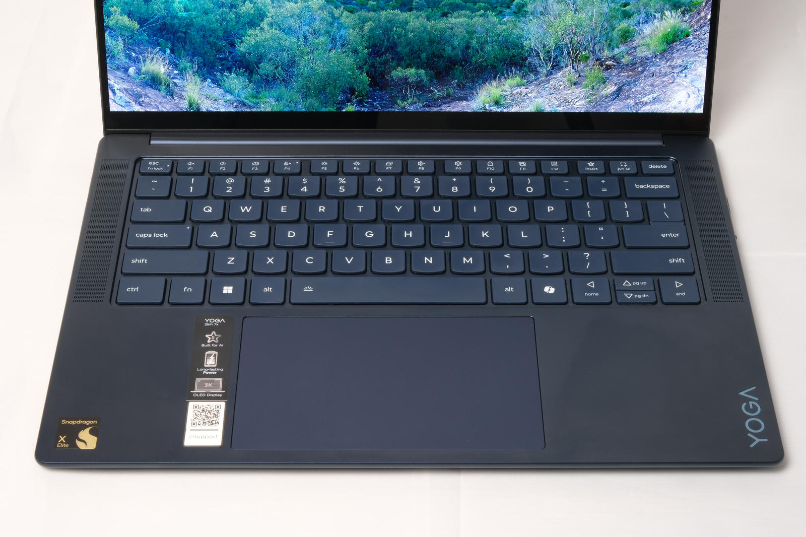 Photo of the keyboard and trackpad. On the right hand palm rest there is a Snapdragon X sticker and a tall sticker from Lenovo.